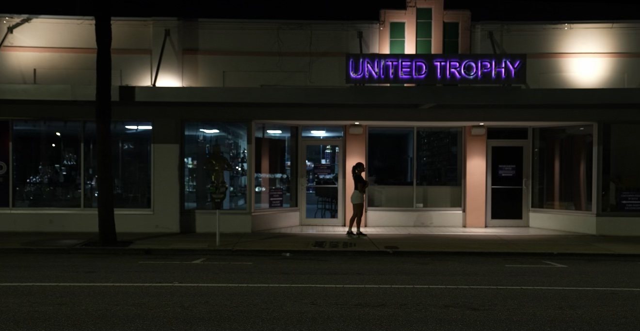 A woman stands alone under a purple neon sign outside a closed storefront at night.