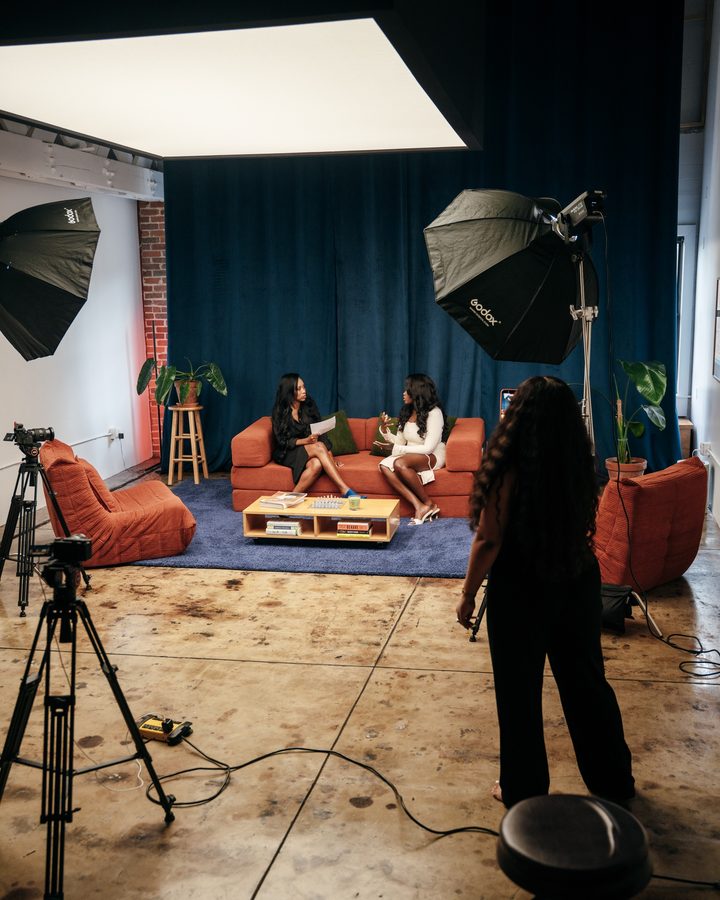 A studio set with two people on a couch under softbox lights, with cameras on tripods in the foreground.